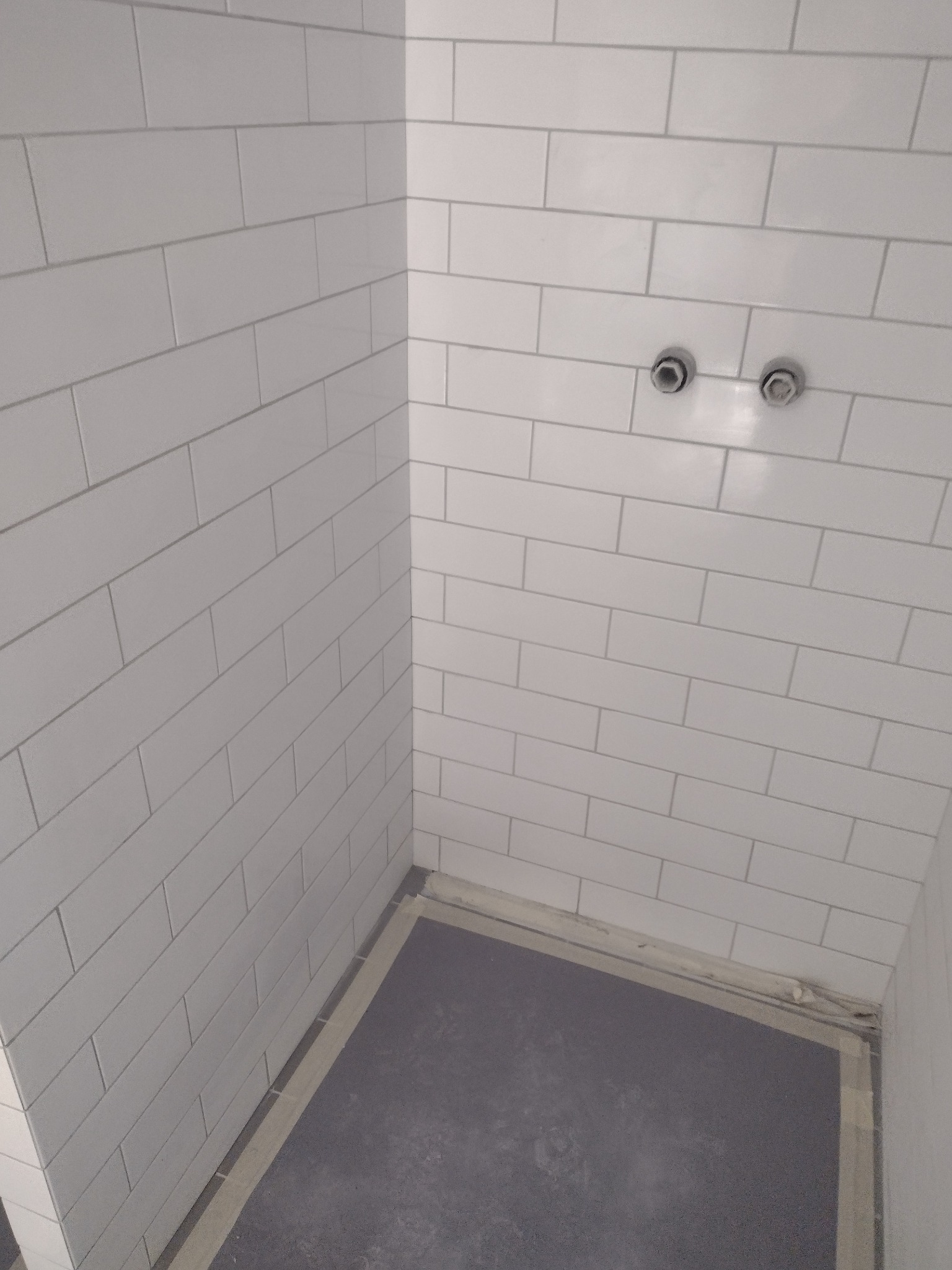 Świeżo ułożone, białe, prostokątne płytki na ścianach w rogu łazienki, z widocznymi dwoma wystającymi przyłączami wodnymi i szarym podkładem pod posadzkę.