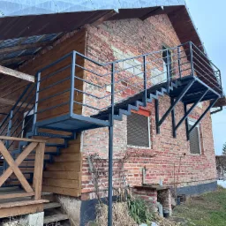 Zewnętrzne metalowe schody prowadzące na balkon przy ceglanym budynku z drewnianą dobudówką, widoczne pozostałości śniegu na trawie.