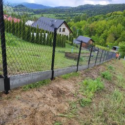 SEMAX Andrzej Sech - Nowoczesne ogrodzenie z siatki z podmurówką betonową na nierównym terenie, w tle dom i zalesione wzgórza. Widoczny fragment bramy i pracownik w odblaskowej kamizelce.