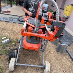 SEMAX Andrzej Sech - Pracownik przy pomarańczowej przecinarce do kostki brukowej na budowie, obok ułożona kostka w szarym odcieniu. Widoczna skrzynka gazowa.