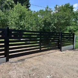 Czarna, przesuwna brama wjazdowa z poziomymi szczeblami, zamontowana na betonowej podstawie. W tle zieleń drzew i fragment ogrodzenia z siatki.