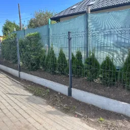 Panelowe ogrodzenie z podmurówką i siatką maskującą, obsadzone tujami. Widok z chodnika, w tle dom z ciemną dachówką i drogowskaz.