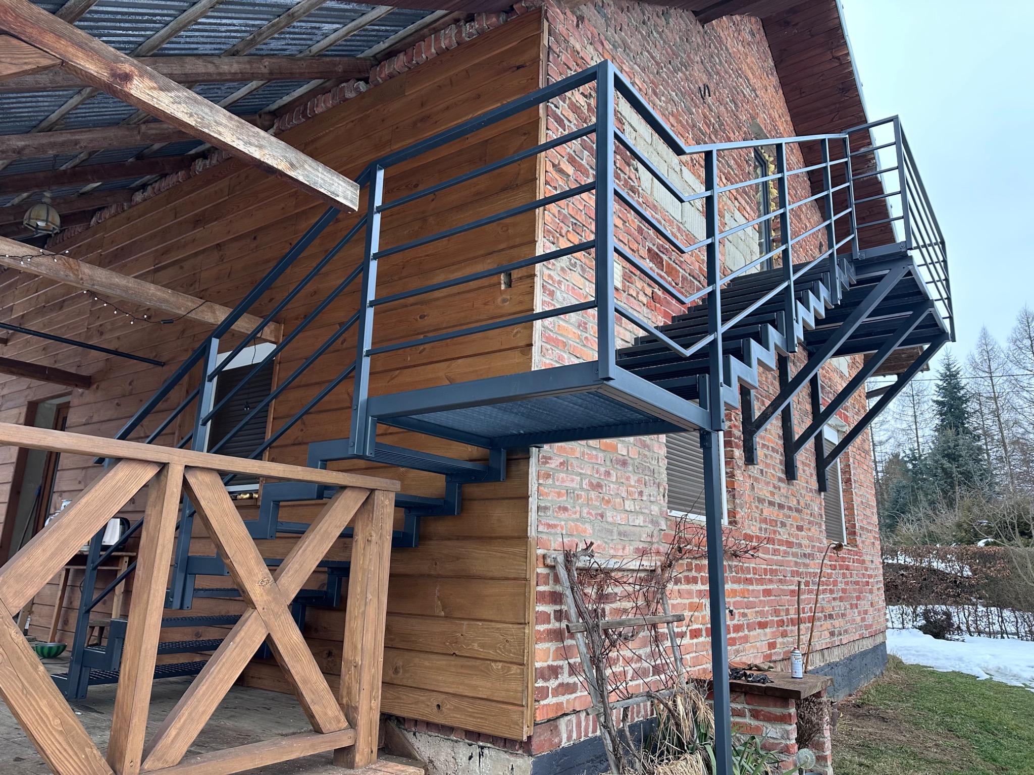 Zewnętrzne schody metalowe z czarną konstrukcją, prowadzące na drewniany taras, zamontowane przy budynku z czerwonej cegły i drewnianą elewacją, widoczne elementy ogrodu w zimowej scenerii.