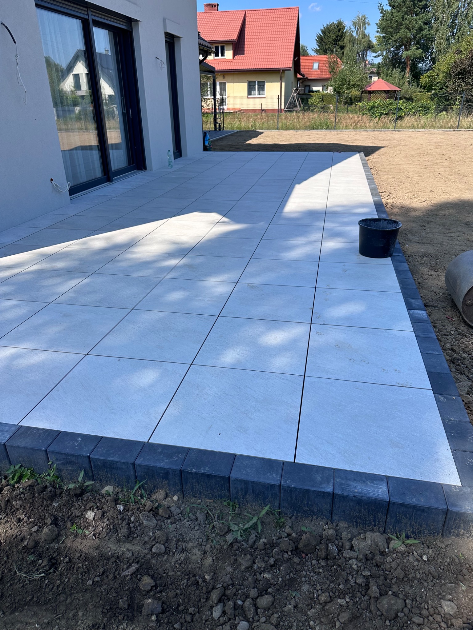 Jasnoszara kostka brukowa ułożona na tarasie przy domu jednorodzinnym. Obramowanie z ciemnej kostki. W tle widoczny ogród i dom sąsiada. Słoneczny dzień.