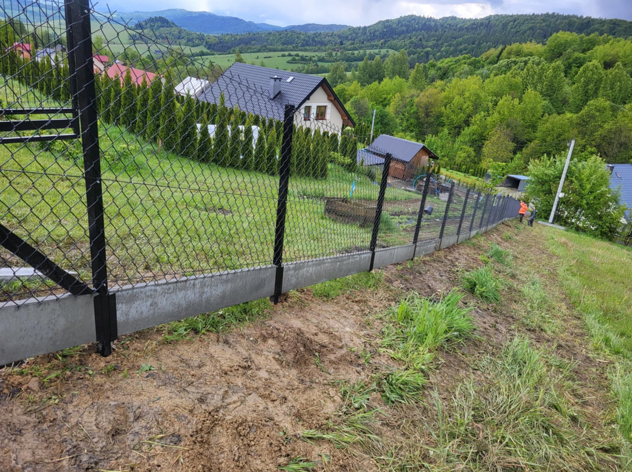 Nowoczesne ogrodzenie z siatki z podmurówką betonową na nierównym terenie, w tle dom i zalesione wzgórza. Widoczny fragment bramy i pracownik w odblaskowej kamizelce.