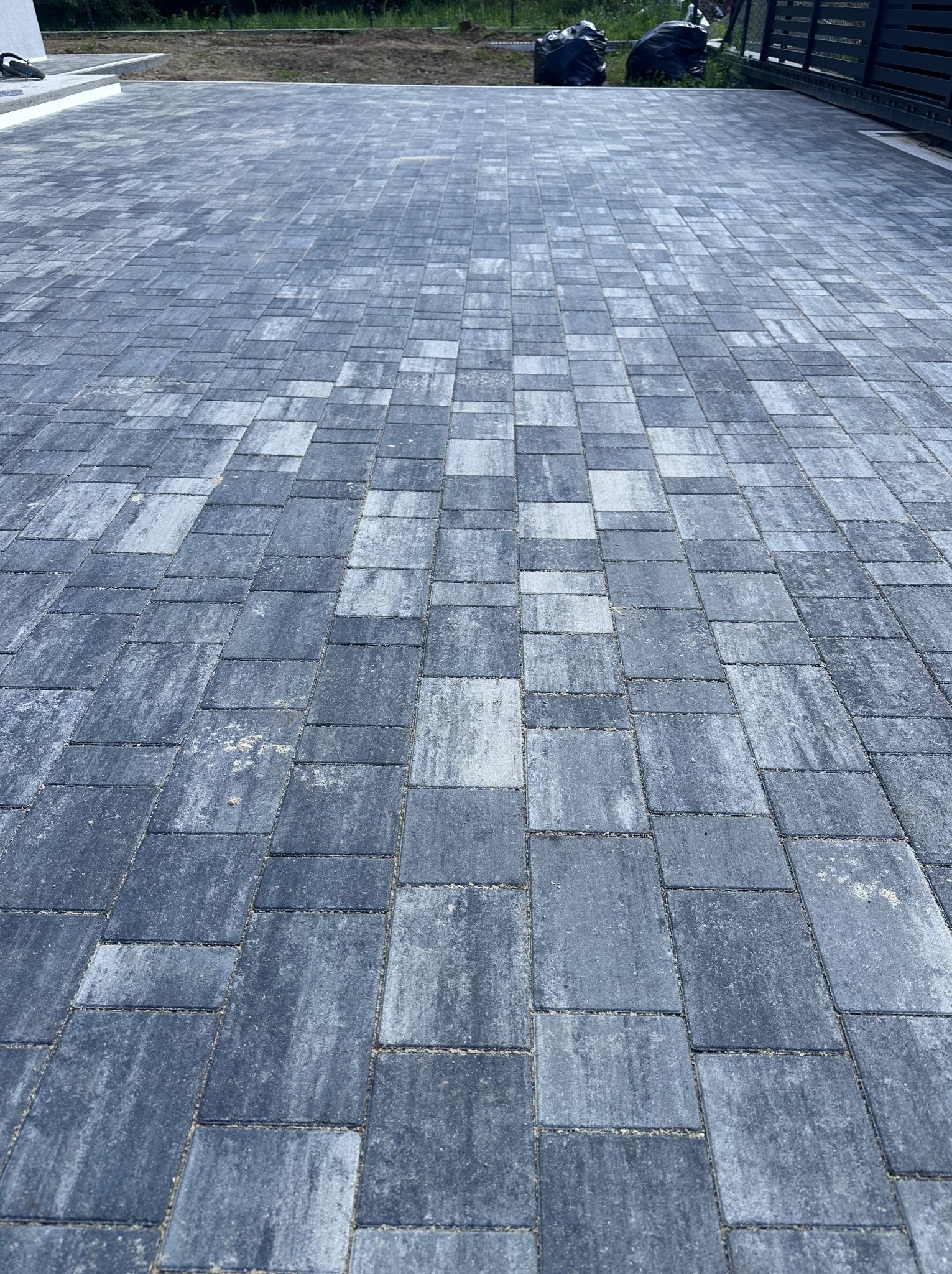 Nowoczesna, szara kostka brukowa ułożona w geometryczny wzór na podjeździe. Widoczne detale faktury kostki i precyzyjne ułożenie. W tle brama wjazdowa i zieleń.