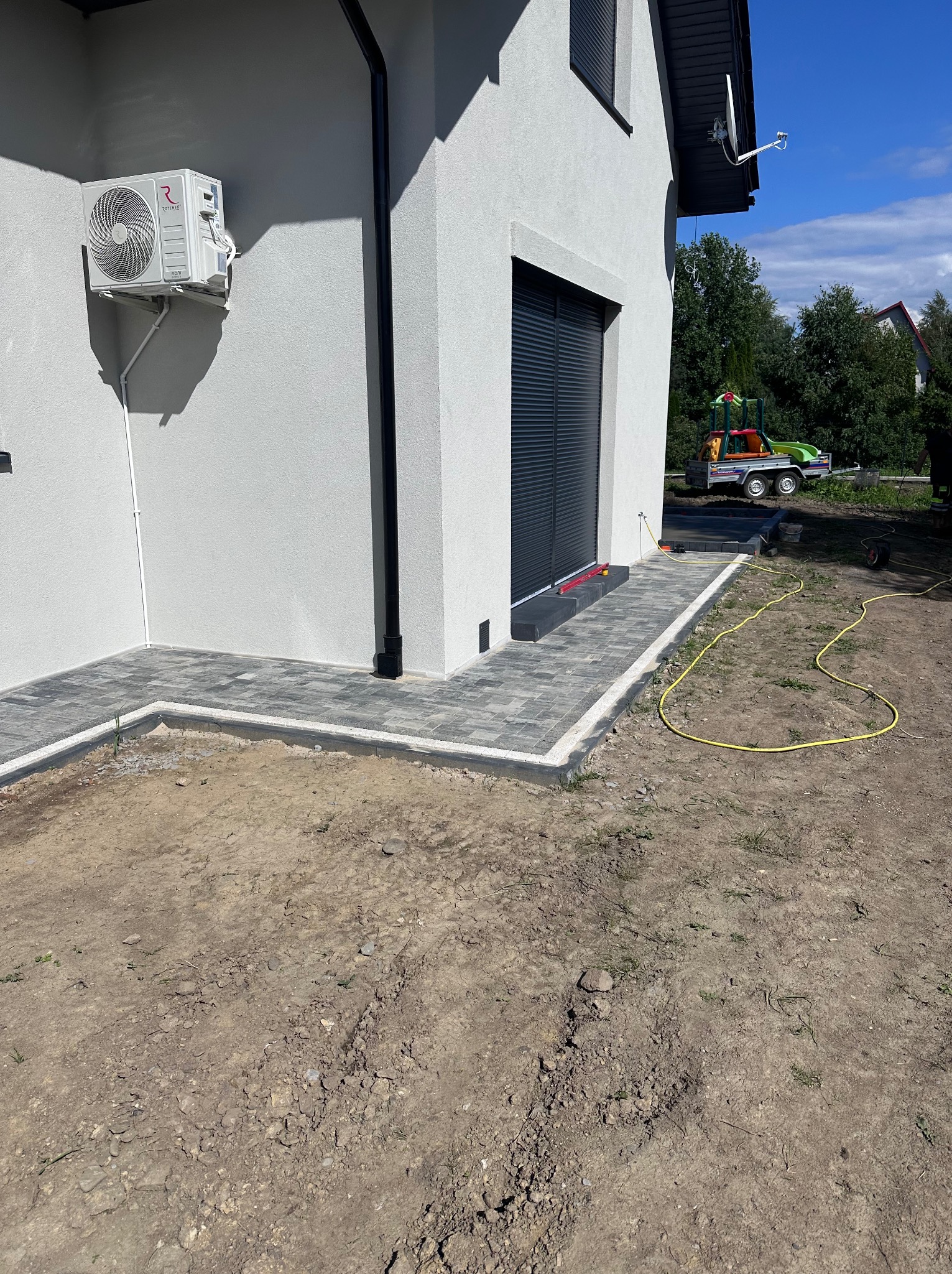 Ułożona kostka brukowa w odcieniach szarości tworzy ścieżkę wokół jasnego budynku z klimatyzatorem. Żółty wąż ogrodowy leży na ziemi. W tle drzewa i plac zabaw.