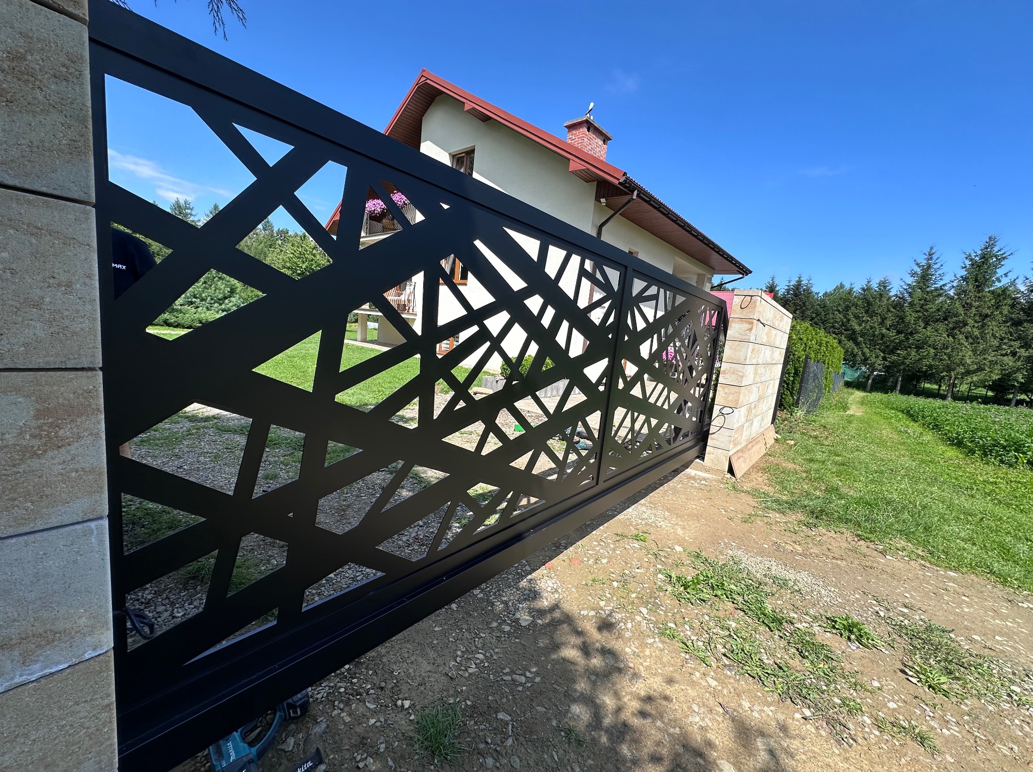 Nowoczesna, czarna brama wjazdowa z ażurowym wzorem geometrycznym, widoczna z boku, na tle domu i zieleni. Elementy montażowe widoczne w dolnej części kadru.