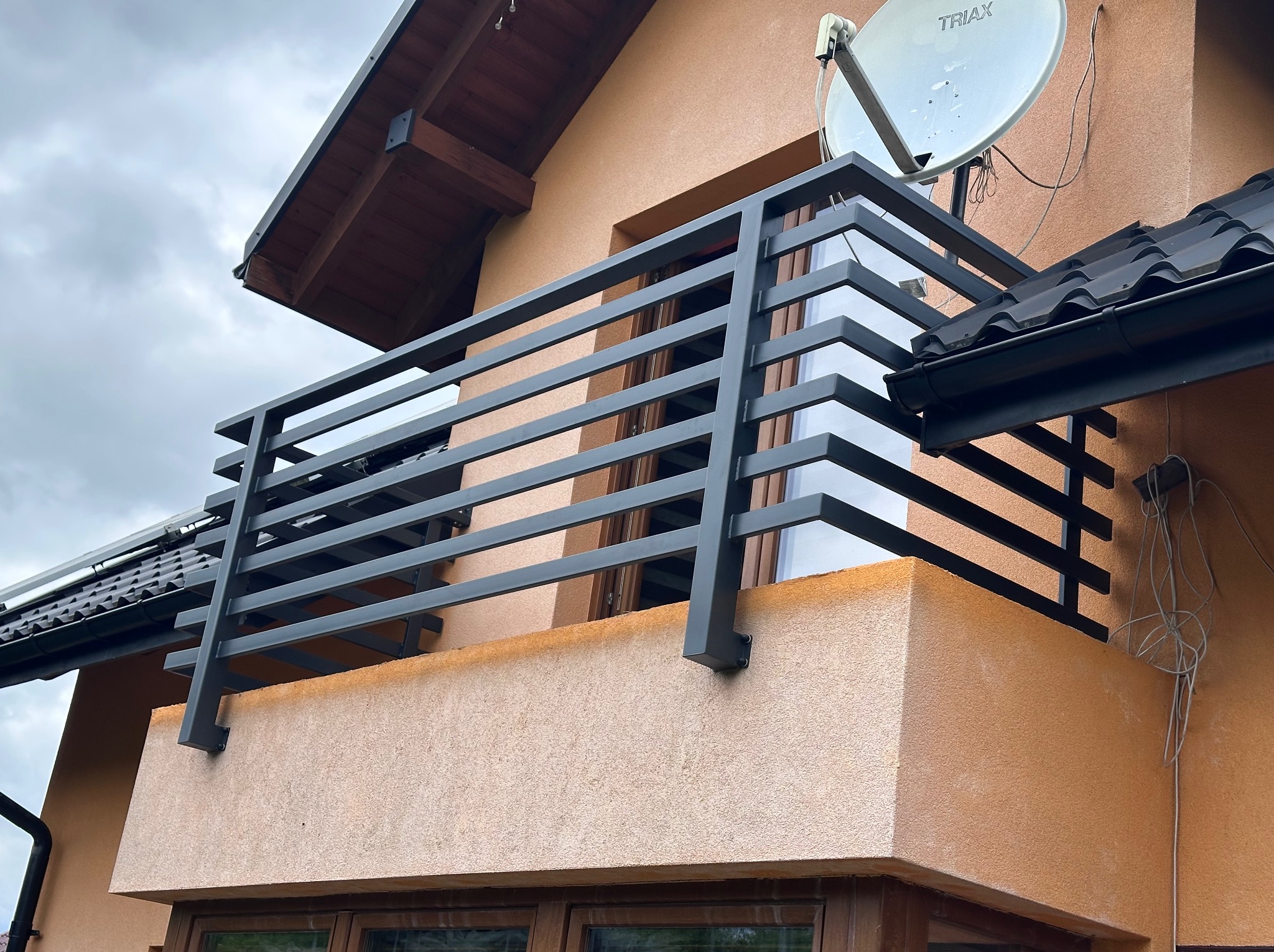 Nowoczesna, pozioma, metalowa balustrada balkonowa w kolorze grafitowym, zamontowana na balkonie budynku o beżowej elewacji, z widoczną anteną satelitarną.