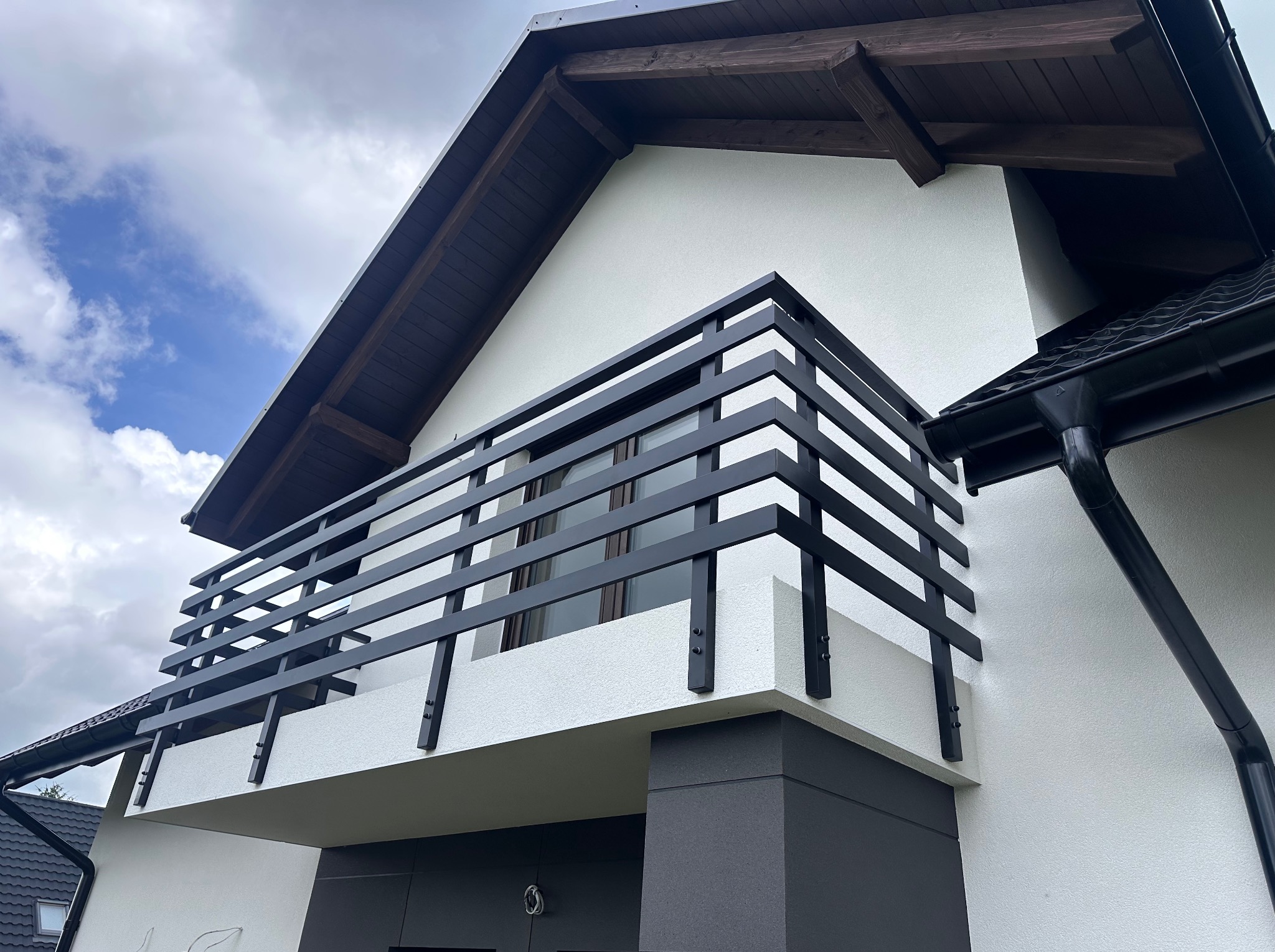 Nowoczesna, czarna balustrada balkonowa z poziomymi szczeblami, zamontowana na białym budynku z elementami szarości. Widok z dołu, pochmurne niebo.