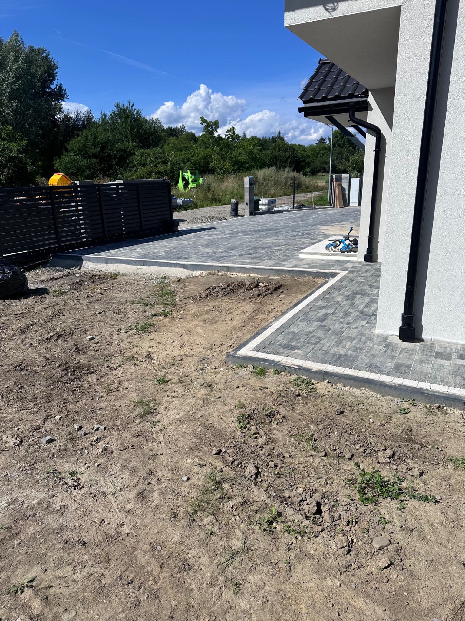 Fragment posesji z ułożoną szarą kostką brukową z obramowaniem. W tle czarne, nowoczesne ogrodzenie panelowe i zieleń. Część terenu jeszcze niezagospodarowana.