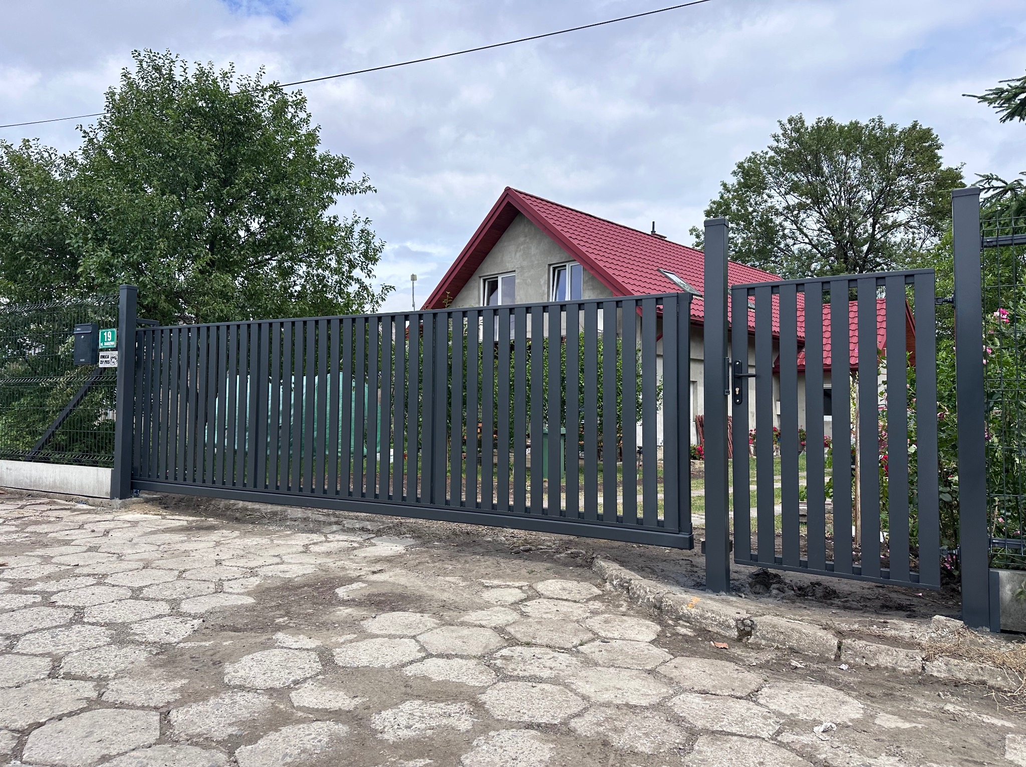 Nowoczesna, przesuwna brama wjazdowa z pionowych profili w kolorze grafitowym, z furtką po prawej stronie. W tle dom z czerwonym dachem i zielone drzewa.
