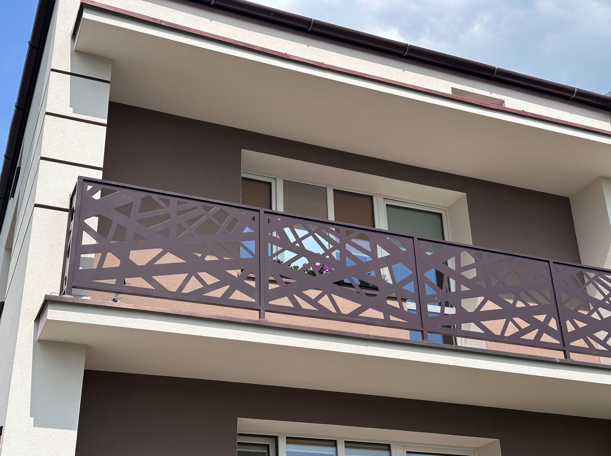 Nowoczesna, ażurowa balustrada balkonowa w geometryczny wzór, brązowa, zamontowana na balkonie budynku z brązowo-białą elewacją. Widok z dołu.