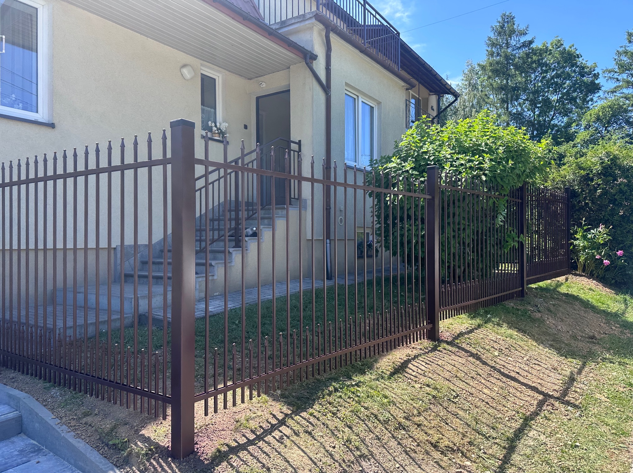 Eleganckie ogrodzenie metalowe w kolorze brązowym, pionowe pręty z ozdobnymi zakończeniami, dopasowane do schodów wejściowych budynku. Trawa i zieleń w tle.