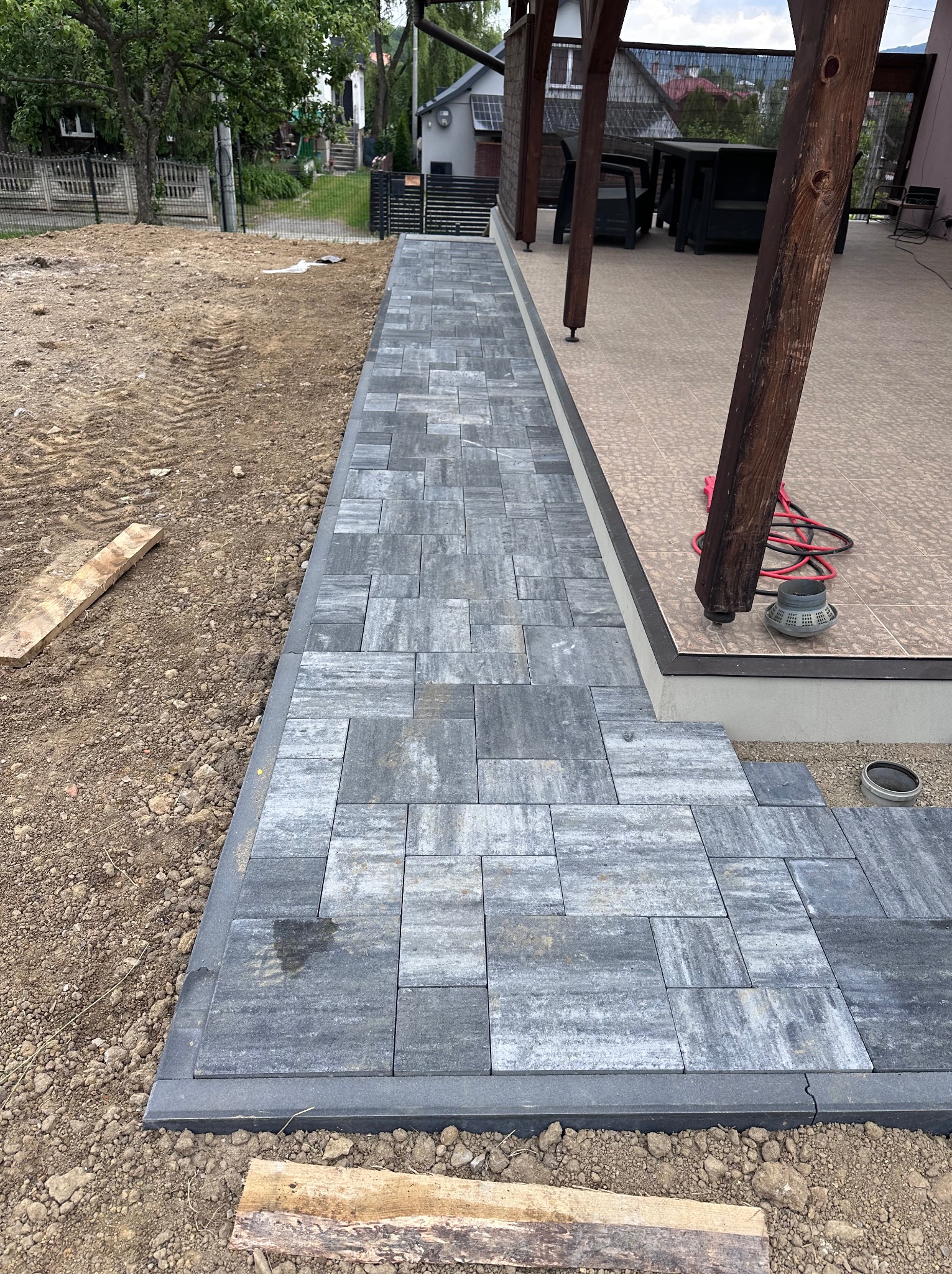Układanie szarej kostki brukowej na ziemi obok tarasu z drewnianymi słupami. Częściowo ułożona nawierzchnia, widoczne narzędzia i materiały budowlane.