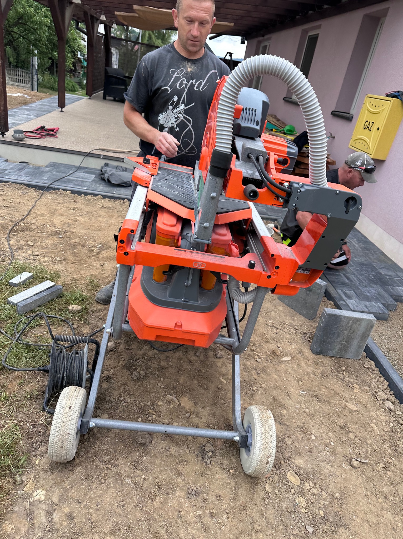 Pracownik przy pomarańczowej przecinarce do kostki brukowej na budowie, obok ułożona kostka w szarym odcieniu. Widoczna skrzynka gazowa.