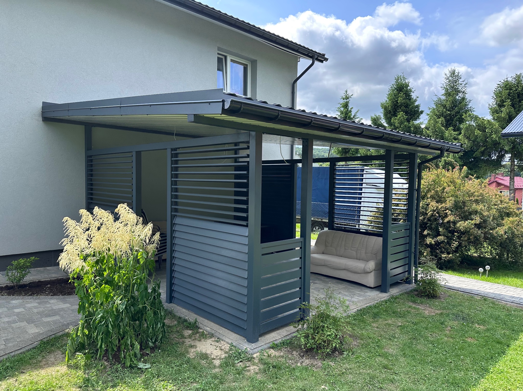 Szara, nowoczesna altana z lamelami i zadaszeniem, przylegająca do budynku, z sofą w środku. Widok na ogród i kostkę brukową.