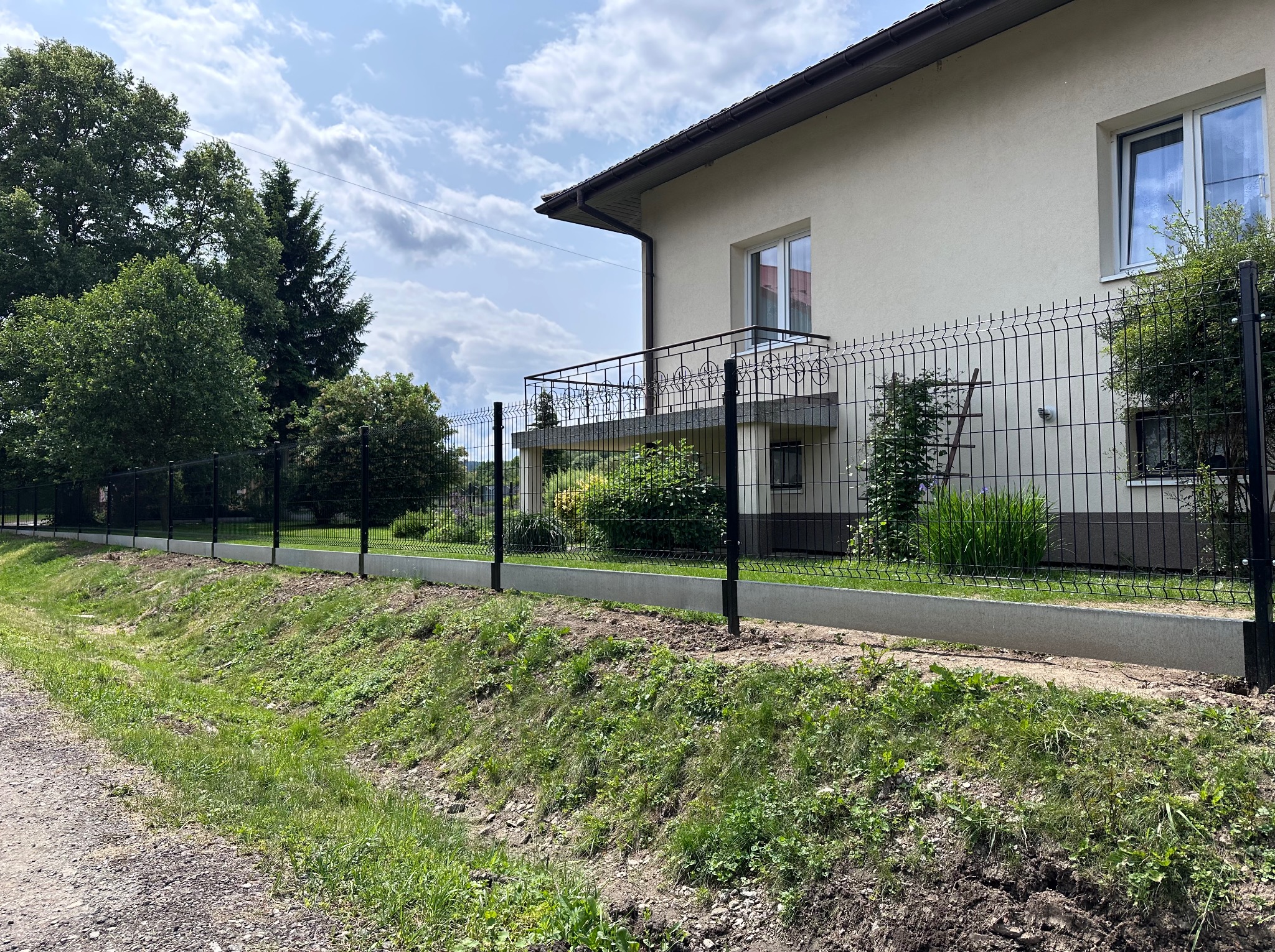 Nowoczesne ogrodzenie panelowe w kolorze grafitowym, zamontowane na betonowej podmurówce, otacza posesję z zielonym trawnikiem i domem w tle.