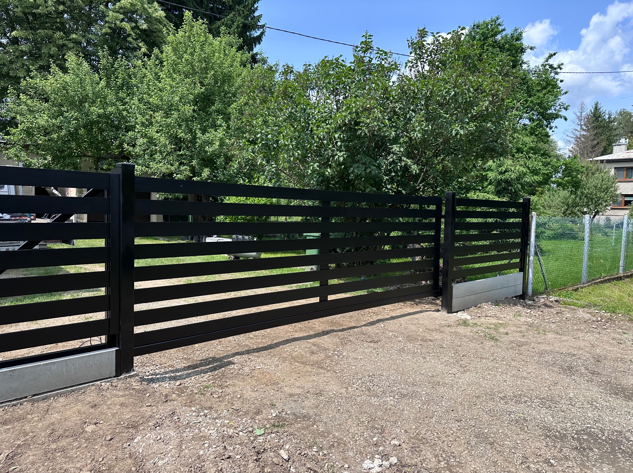 Czarna, przesuwna brama wjazdowa z poziomymi szczeblami, zamontowana na betonowej podstawie. W tle zieleń drzew i fragment ogrodzenia z siatki.