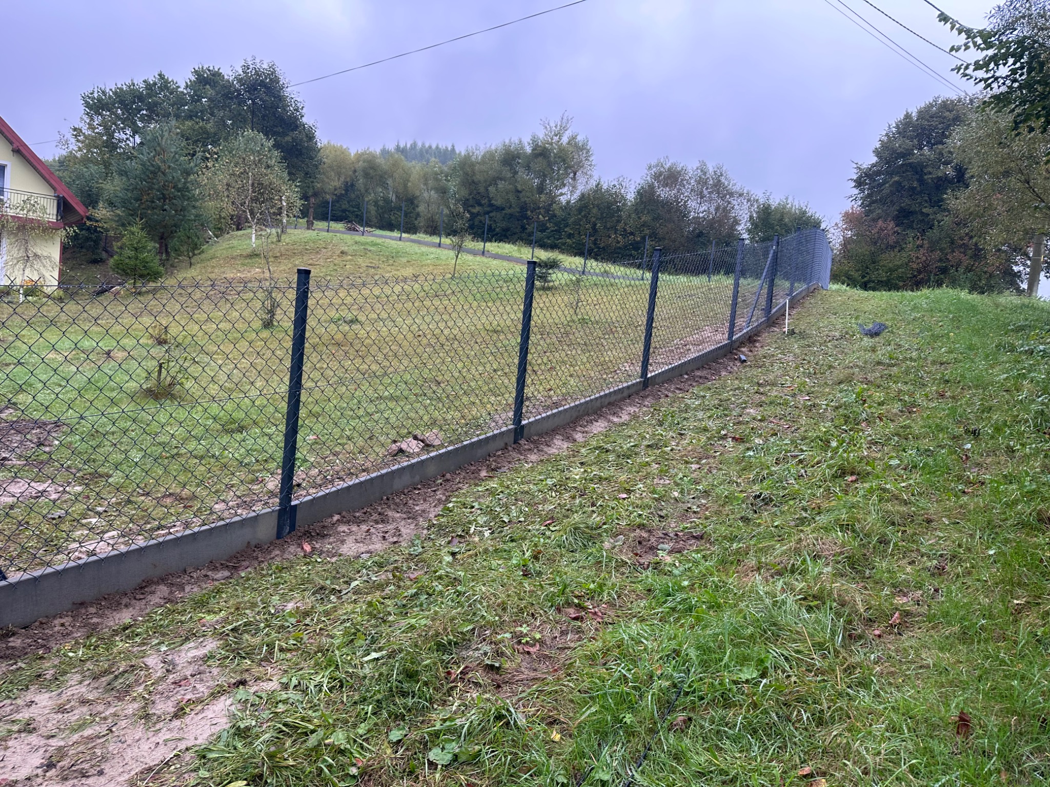 Nowo postawione ogrodzenie z siatki na betonowej podmurówce, biegnące wzdłuż pagórkowatego terenu z widokiem na drzewa i dom w tle. Widoczne słupki i naciągnięta siatka.