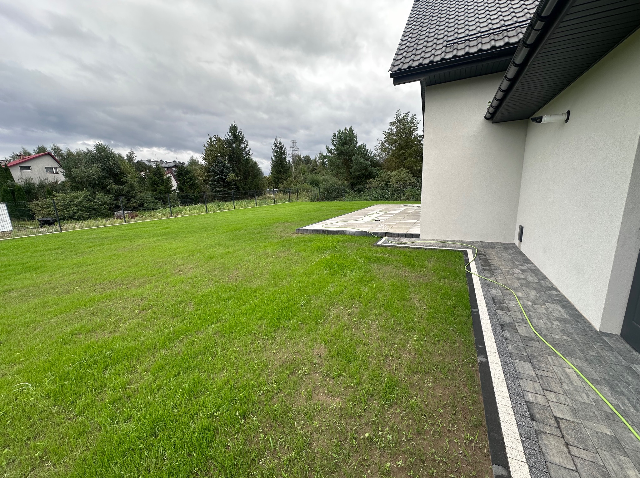 Nowoczesny dom z szarą kostką brukową na ścieżce i tarasie, zielony trawnik, pochmurne niebo. Widok z boku, częściowo widoczny budynek i otoczenie.