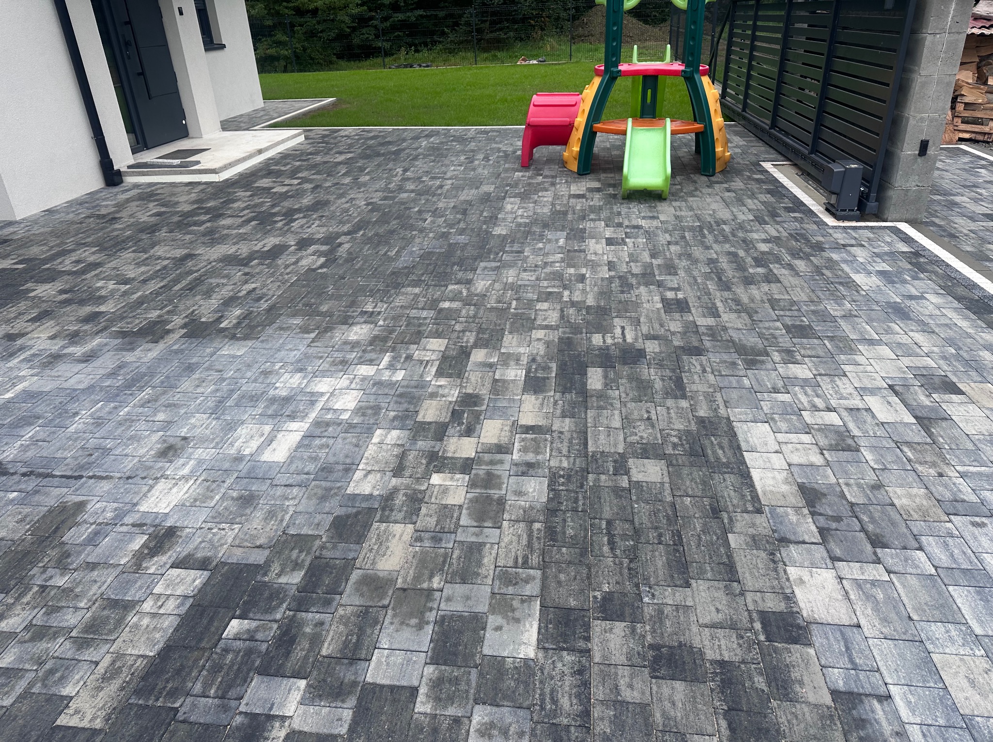 Ułożona szara kostka brukowa na podjeździe, z widocznym fragmentem domu, bramą przesuwną i elementami placu zabaw w tle. Perspektywa z dołu podkreśla wzór kostki.