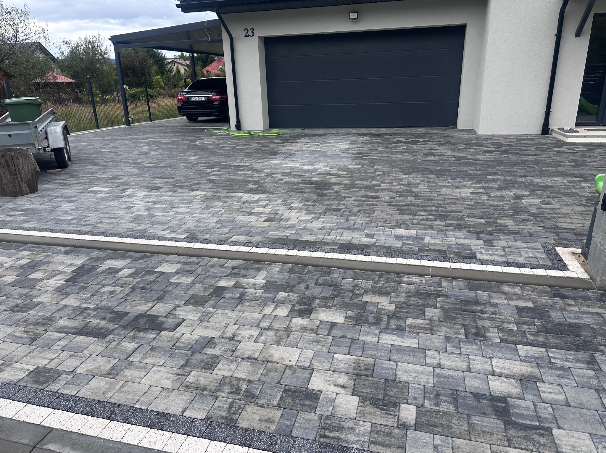 Ułożona kostka brukowa w odcieniach szarości przed nowoczesnym domem z garażem i podjazdem. Widoczne obrzeża i dbałość o detale wykończenia.