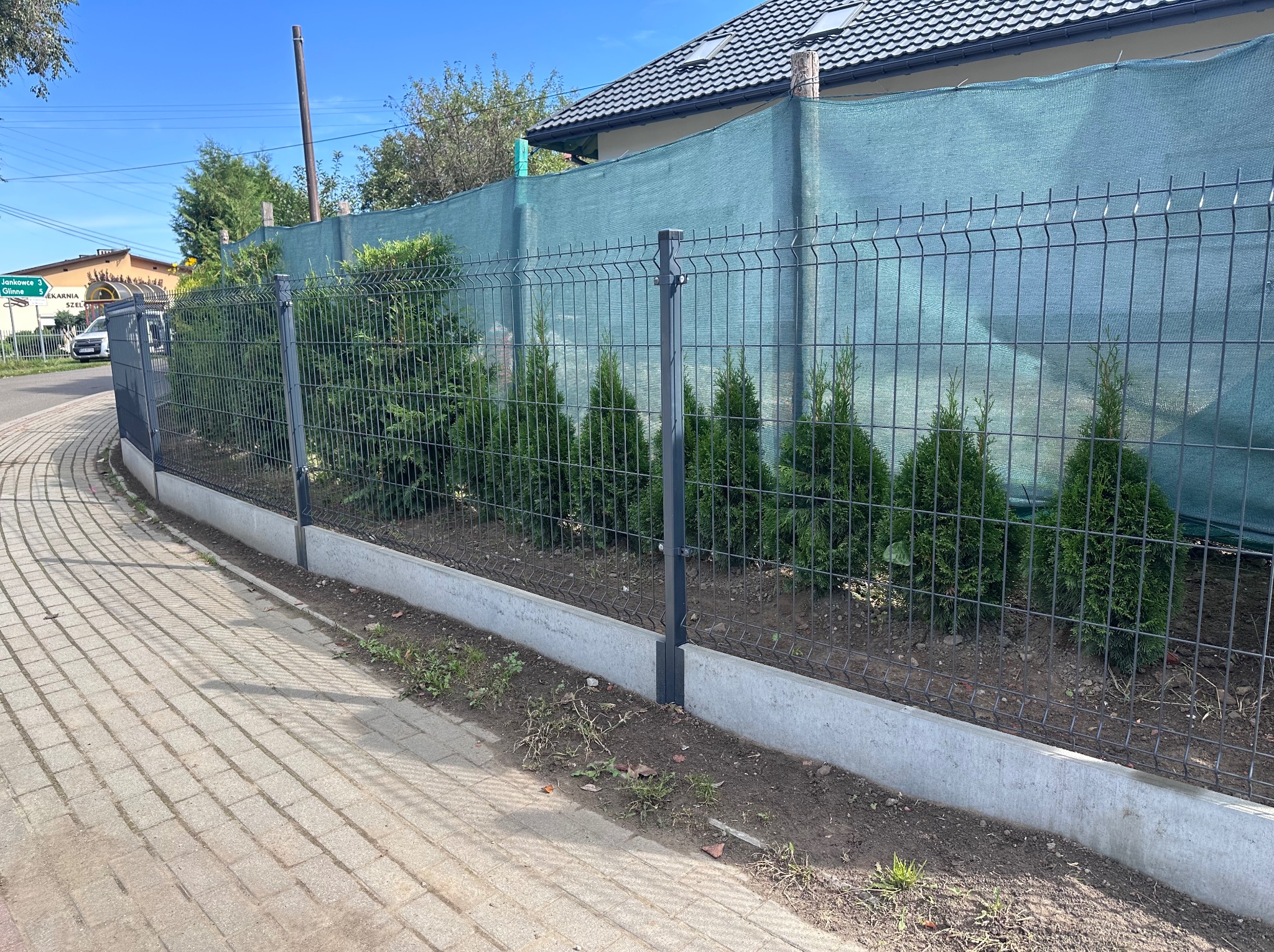 Panelowe ogrodzenie z podmurówką i siatką maskującą, obsadzone tujami. Widok z chodnika, w tle dom z ciemną dachówką i drogowskaz.
