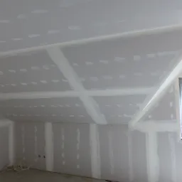 Poddasze z zamontowanymi płytami gipsowo-kartonowymi, widoczne spoiny i szpachlowanie, surowa żarówka zwisająca z sufitu, okno z niebieską taśmą zabezpieczającą.