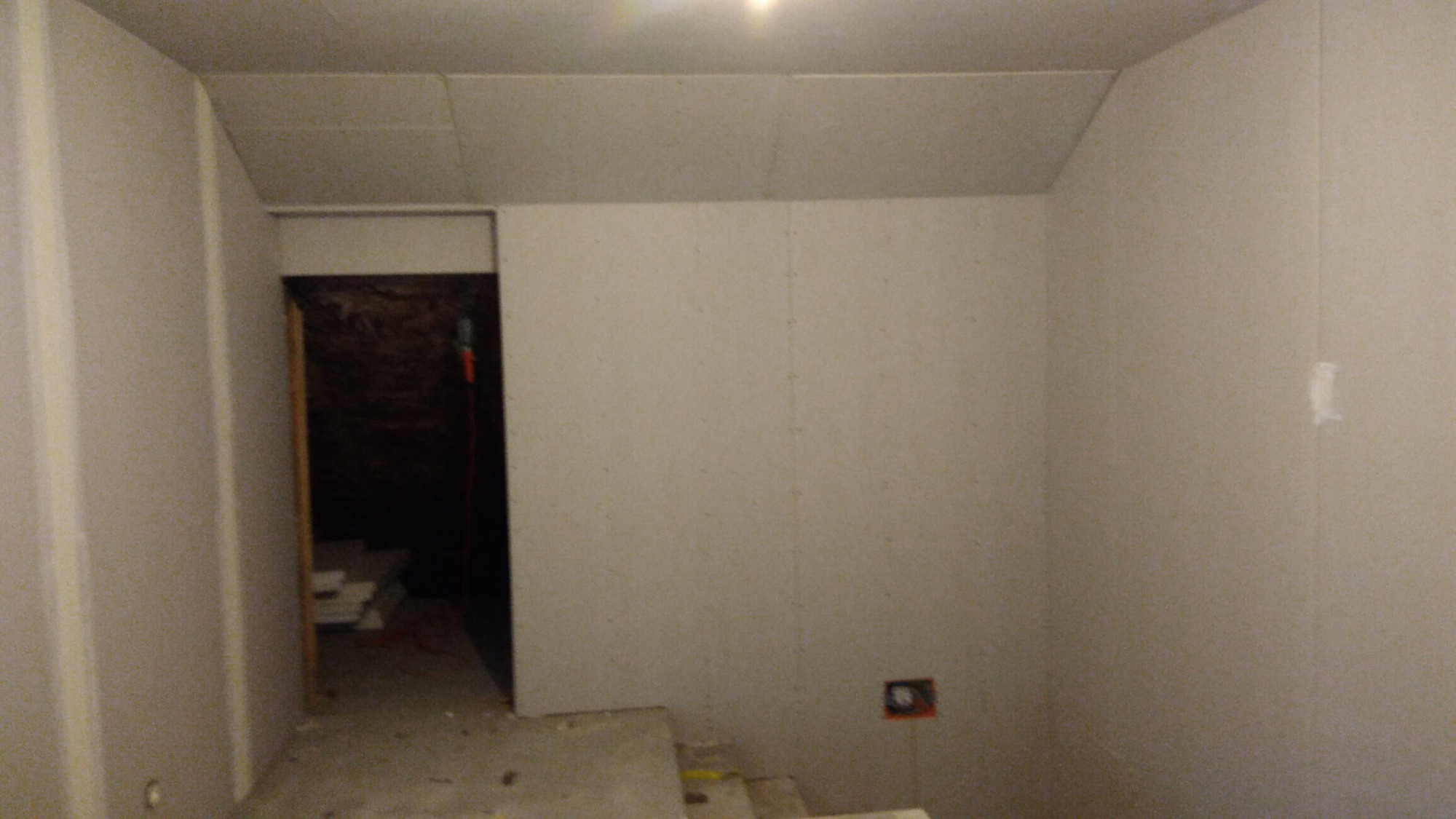 Wnętrze w trakcie wykańczania płytami gipsowo-kartonowymi, widoczne połączenia płyt i otwór drzwiowy z widocznym okablowaniem. Częściowo oświetlone pomieszczenie z surowym betonem na podłodze.