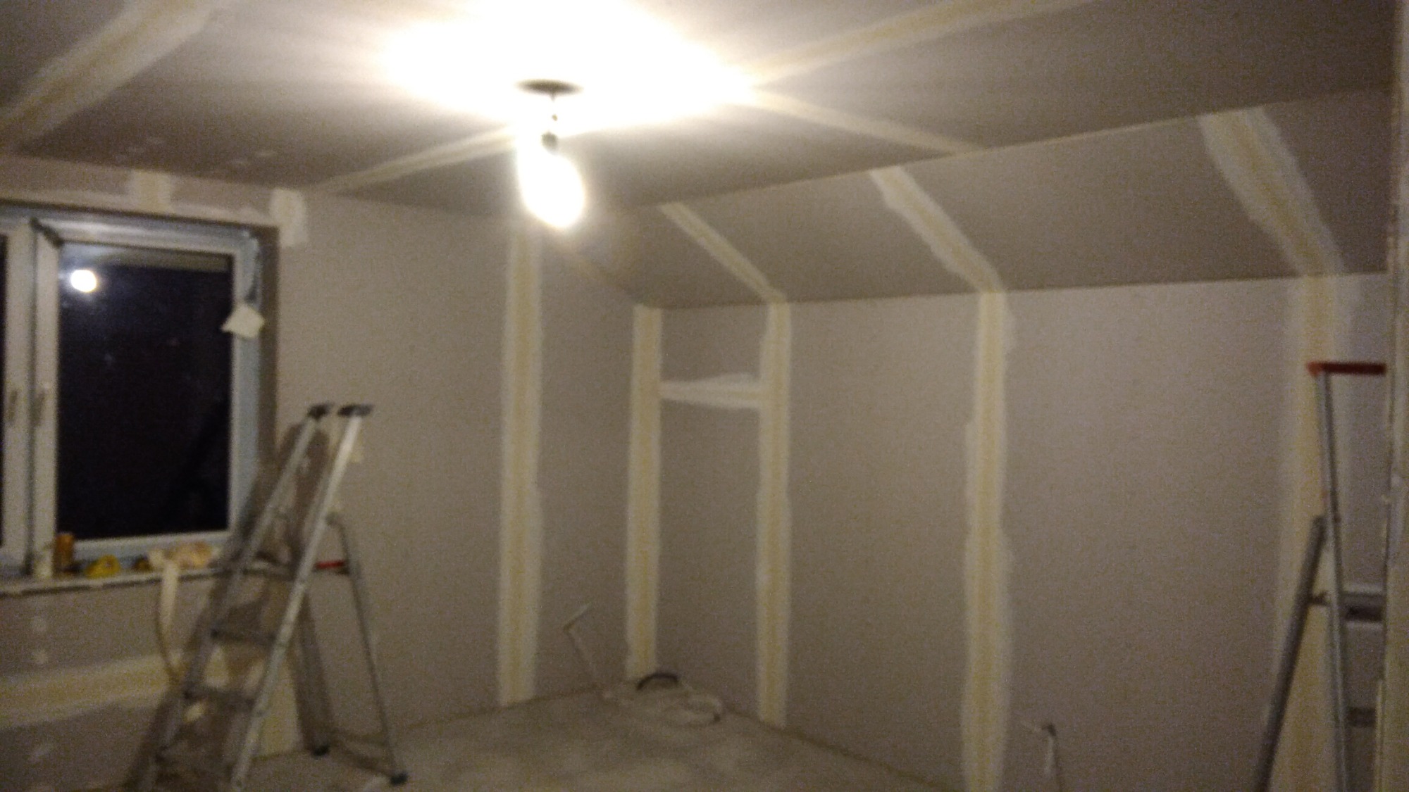 Poddasze w trakcie prac wykończeniowych, ściany pokryte płytami gipsowo-kartonowymi z widocznymi łączeniami wzmocnionymi taśmą, drabina przystawiona do ściany obok okna, surowa żarówka oświetla...