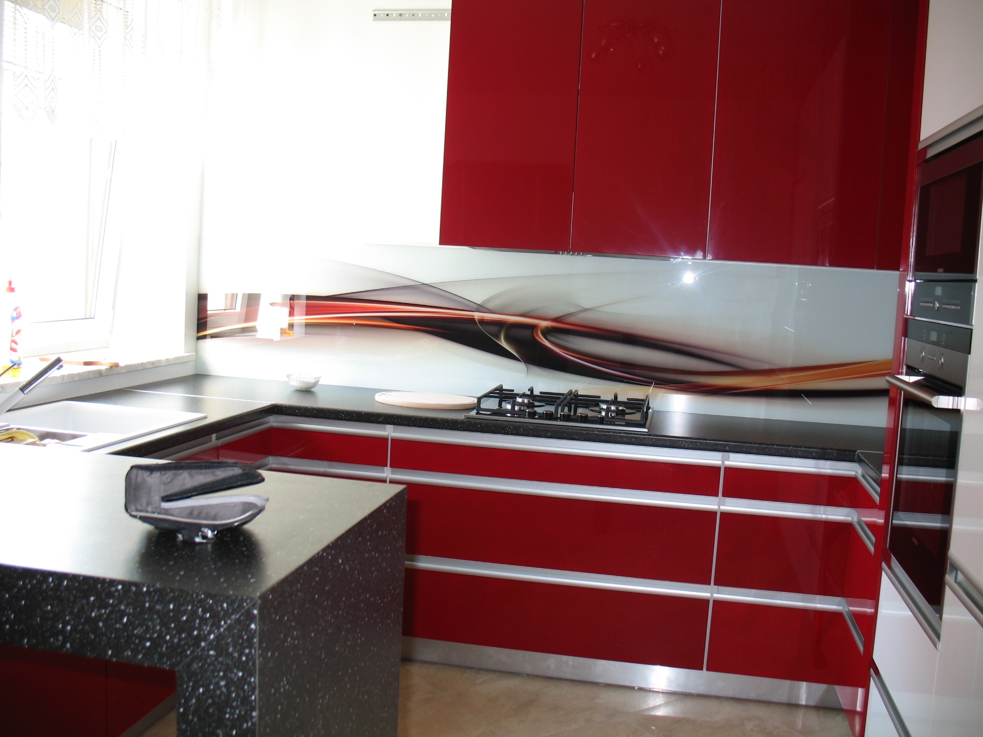 Nowoczesna kuchnia z czerwonymi, lakierowanymi frontami szafek, czarnym blatem z nakrapianym wzorem i szklanym panelem dekoracyjnym nad blatem.