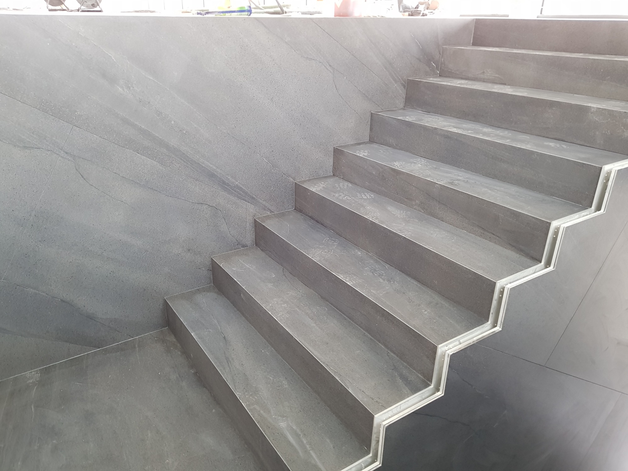 Wykończone schody wewnętrzne z szarej płytki imitującej kamień, z metalowymi listwami krawędziowymi, widoczne ślady montażu.