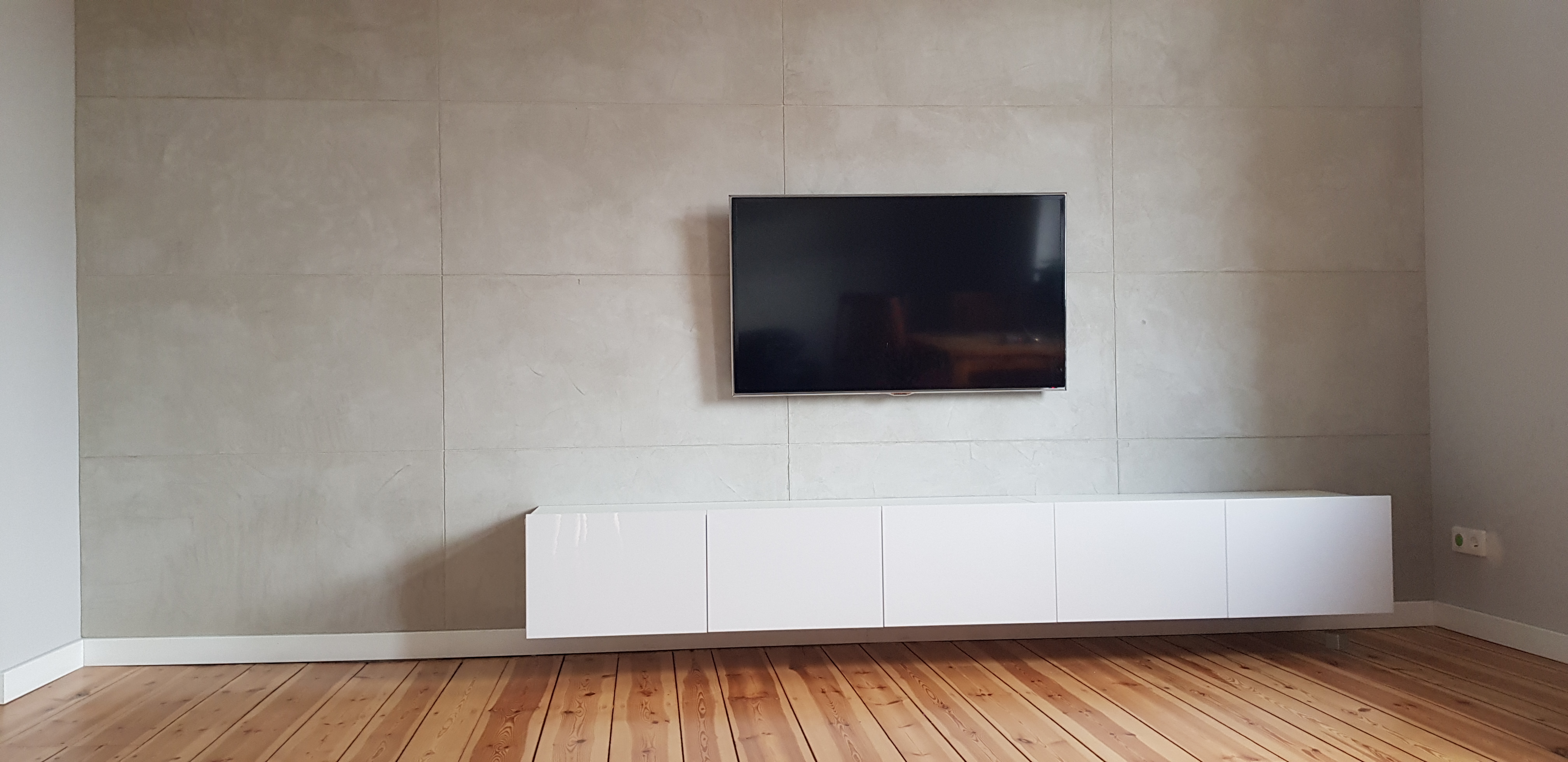 Ściana wykończona betonem architektonicznym w jasnych odcieniach szarości, wiszący telewizor i biała, podwieszana szafka RTV, drewniana podłoga z widocznymi słojami.