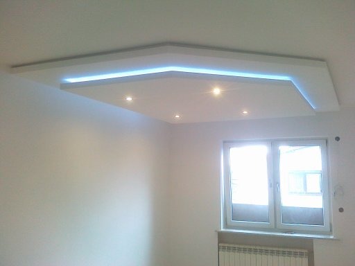 Nowoczesny sufit podwieszany z oświetleniem LED w kształcie geometrycznym, z widocznymi punktowymi lampami i oknem w tle.