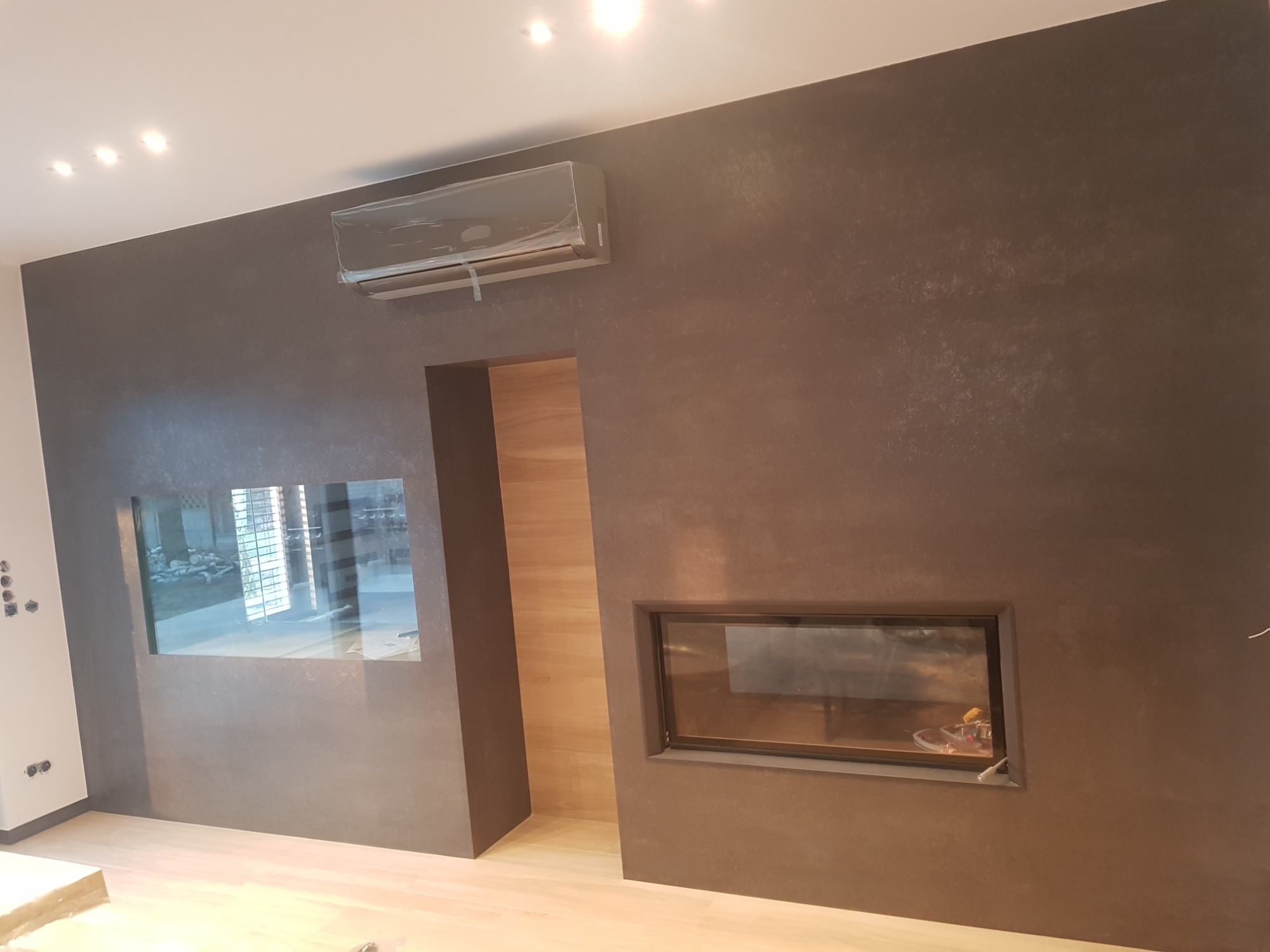 Strukturalna ściana w odcieniach brązu z wbudowanym akwarium i kominkiem, klimatyzator zabezpieczony folią, jasna drewniana podłoga.