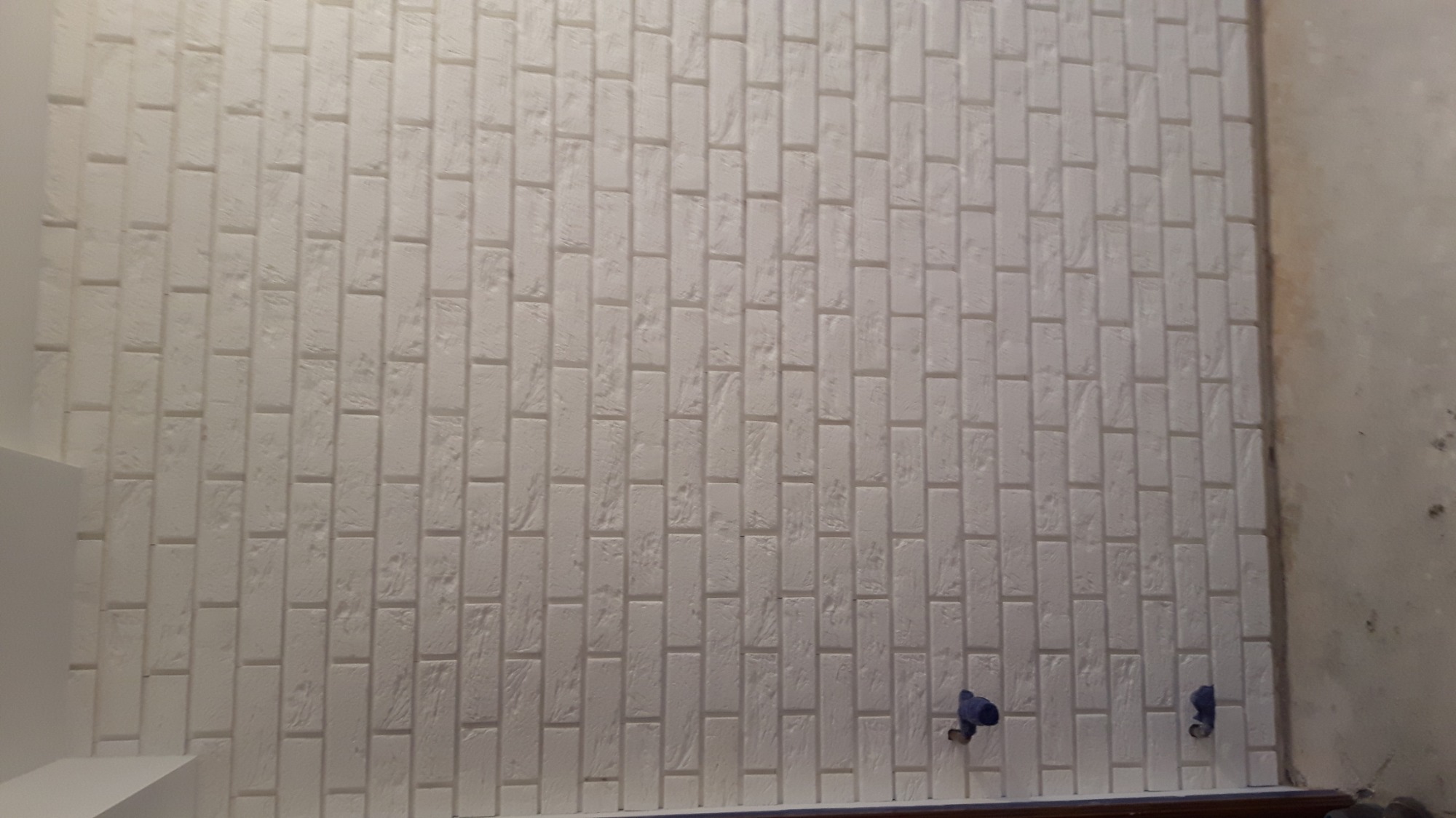 Biała ściana wykończona płytkami imitującymi cegłę, widoczne instalacje hydrauliczne w trakcie prac.