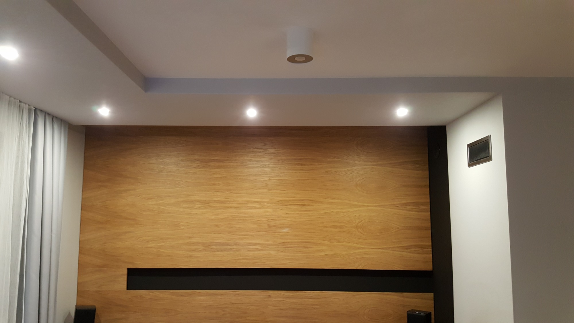 Ściana z drewnianą okładziną z czarnym wnęką, oświetlona punktowymi lampami sufitowymi i zasłoniętym oknem po lewej stronie. Widoczny fragment ściany z gniazdkiem.