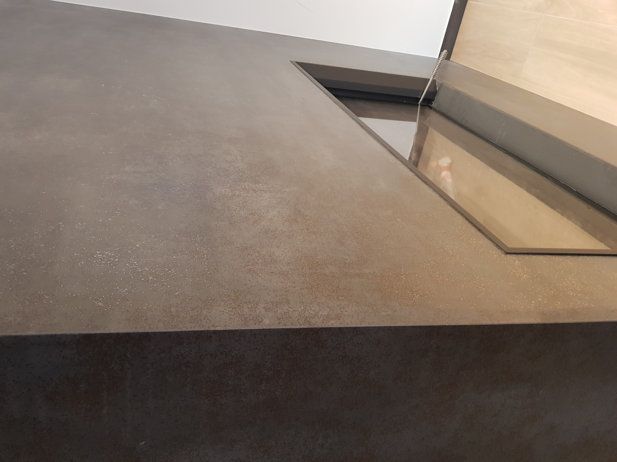 Szara, teksturowana ściana z trapezoidalnym oknem w górnej części, widok z dołu, ukazujący nieregularną fakturę i subtelne refleksy świetlne.