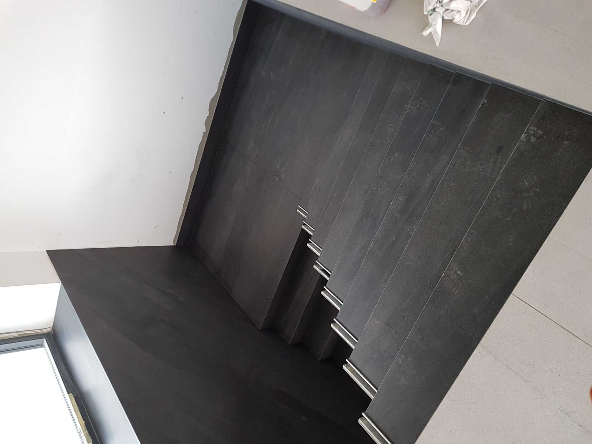 Wykończone schody z ciemnych, grafitowych płytek z metalowymi listwami krawędziowymi, widok z góry, częściowo widoczna biała ściana i okno.