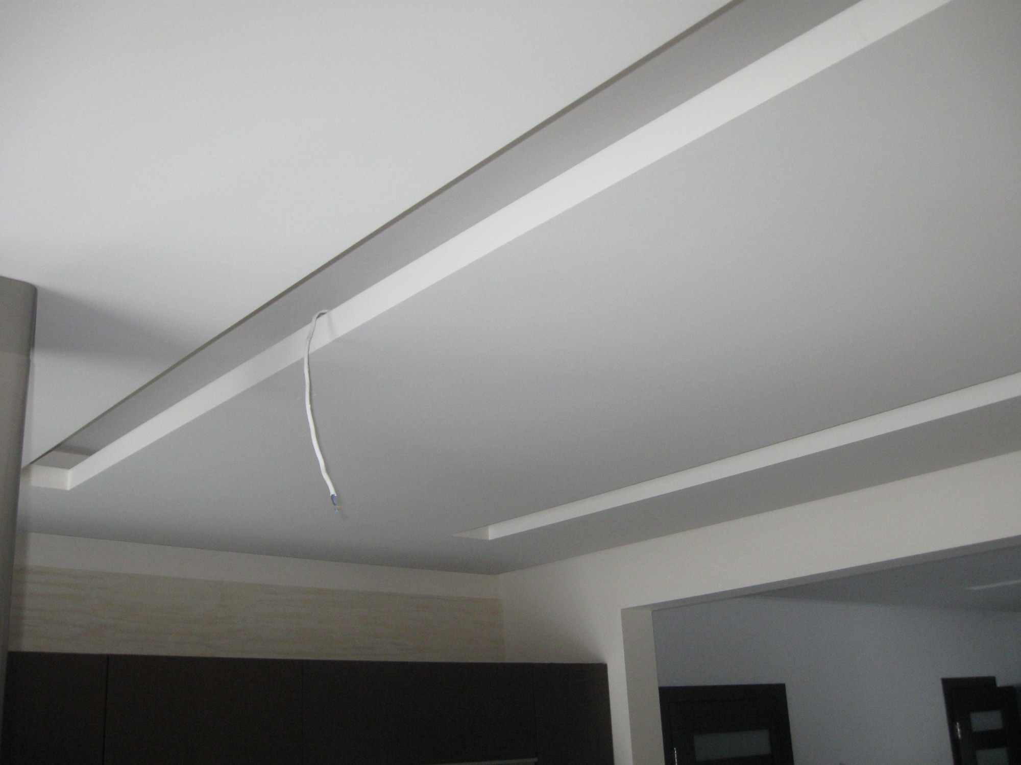 Biały sufit napinany z dekoracyjnymi listwami i wystającym kablem elektrycznym w rogu pomieszczenia.
