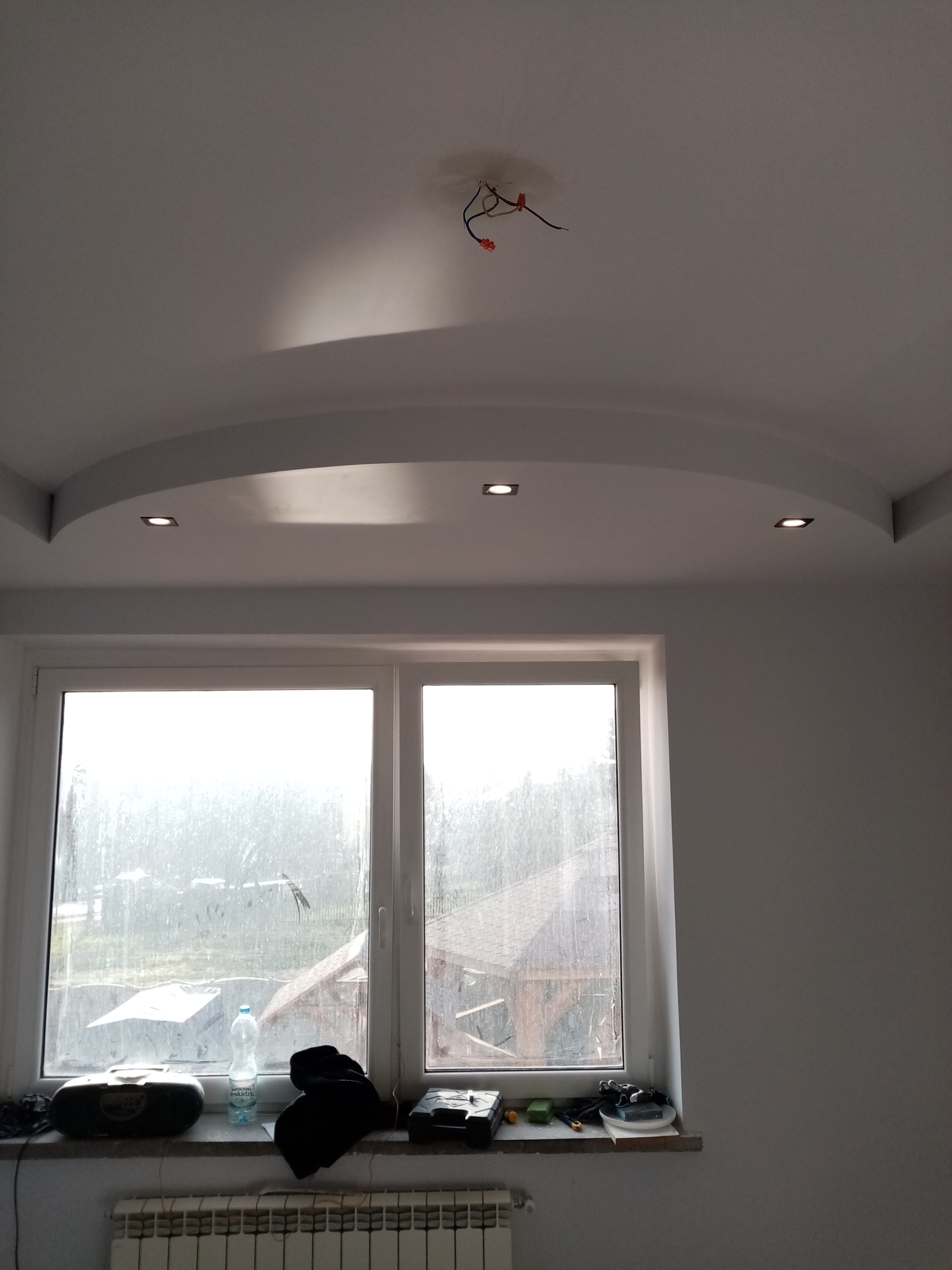 Podwieszany sufit w łazience z wbudowanymi punktowymi lampami, widoczne kable do podłączenia oświetlenia centralnego, okno z widokiem na zewnątrz.