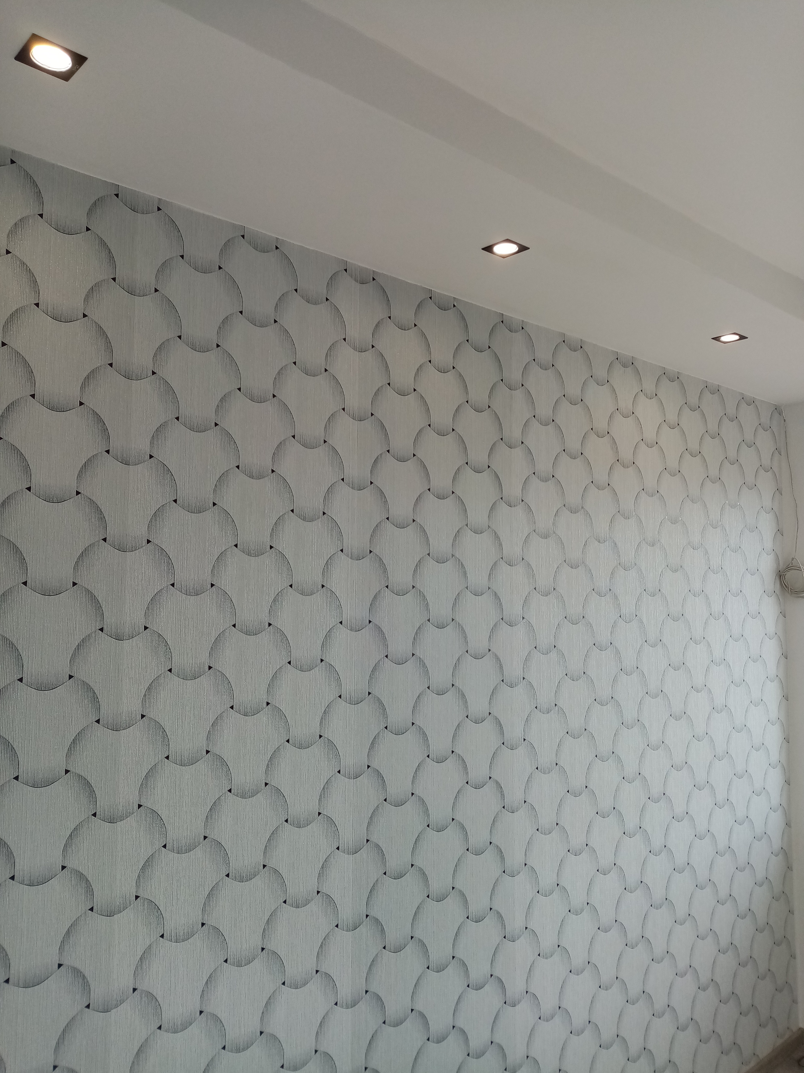 Ściana w łazience wykończona tapetą z motywem szarych półkoli i czarnych trójkątów, oświetlona trzema kwadratowymi lampami sufitowymi.