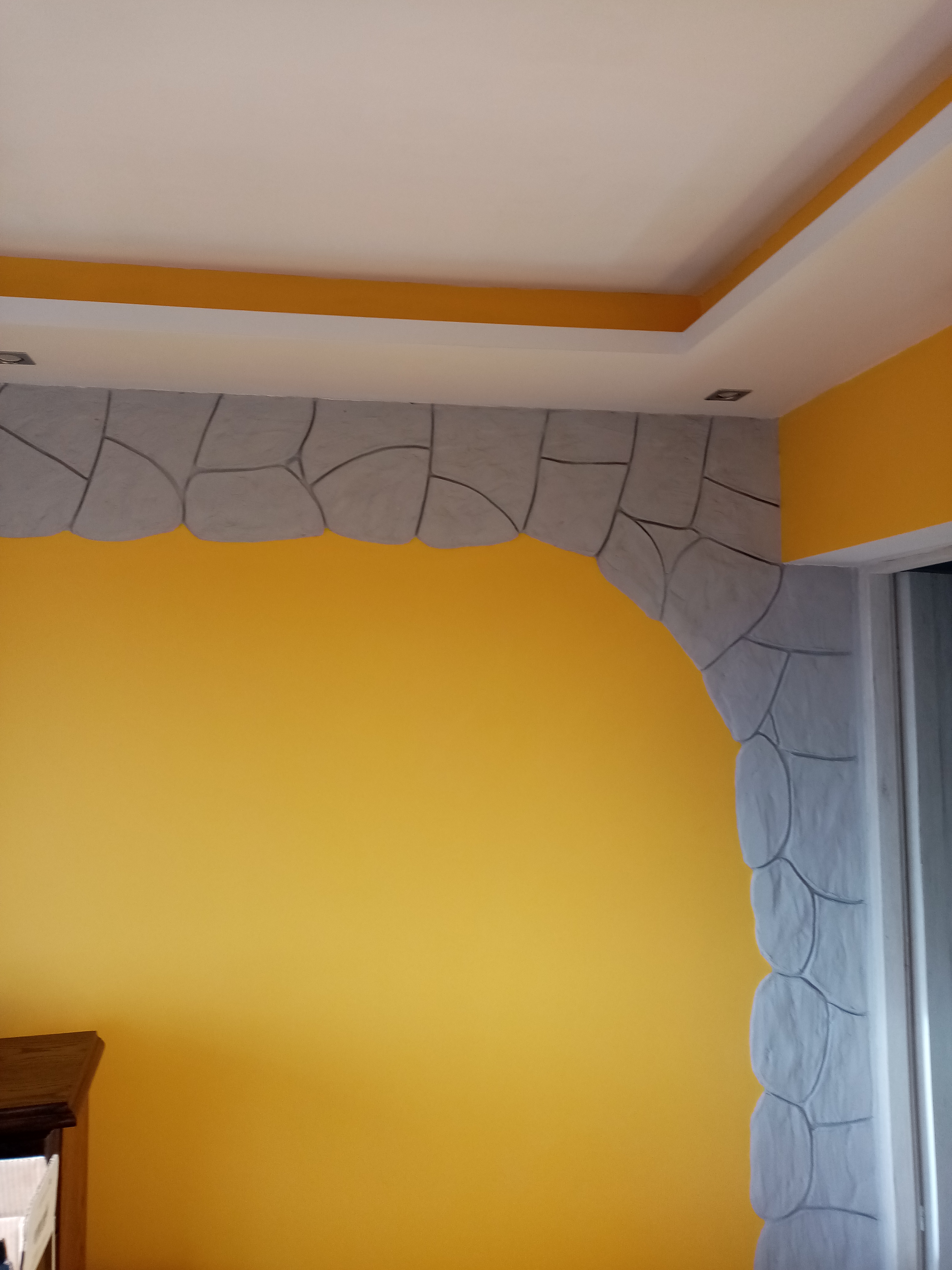 Żółta ściana z dekoracyjnym łukiem z imitacji kamienia w szarym kolorze, oświetlenie sufitowe.