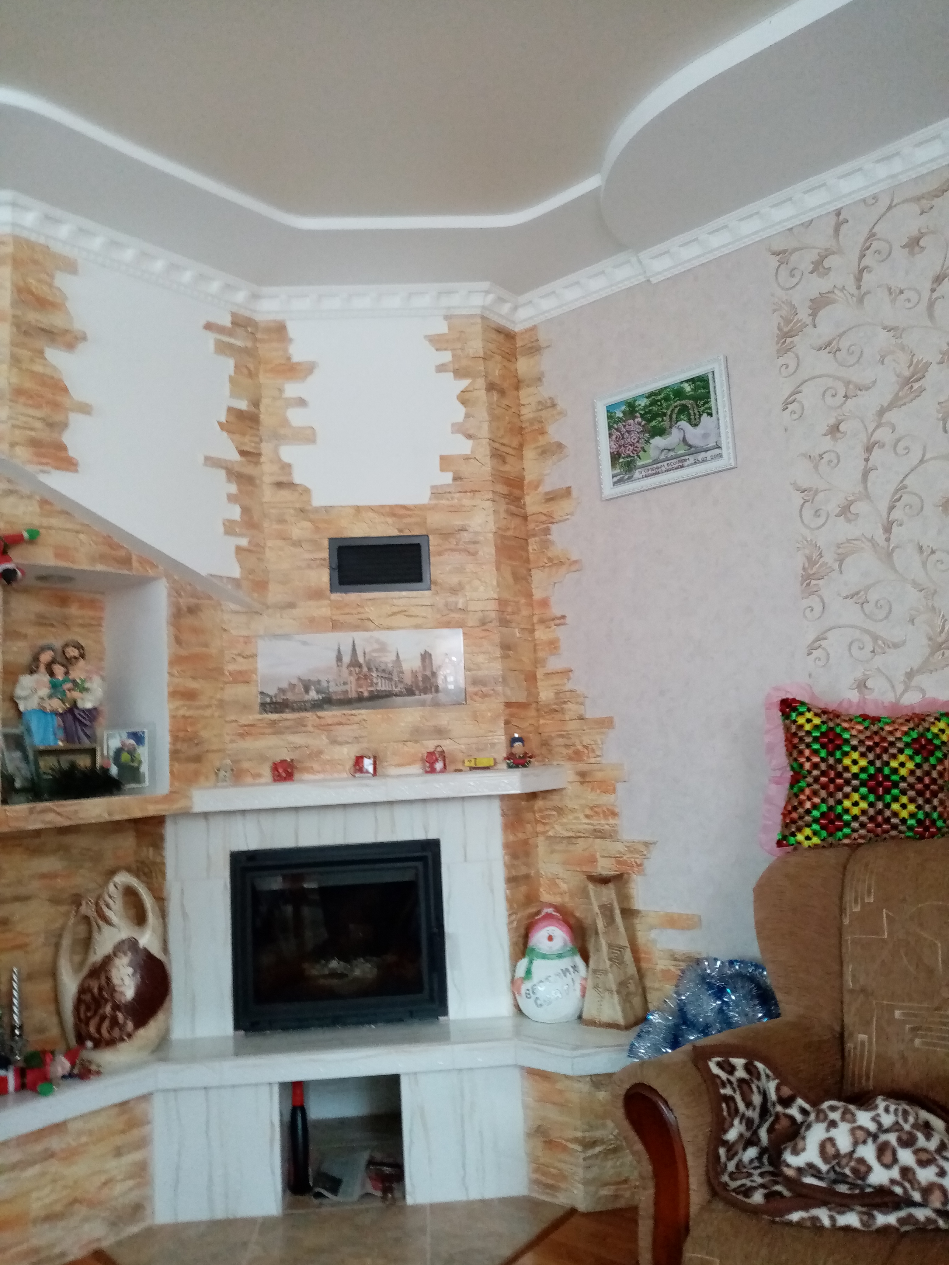 Wykończony narożny kominek obłożony cegłą dekoracyjną, z półkami ozdobionymi figurkami i obrazami, widoczny fragment fotela z poduszką i kocem w panterkę.