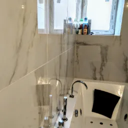 Biała wanna z hydromasażem, chromowana armatura, czarna poduszka, marmurowe płytki na ścianie, okno z widocznymi kosmetykami.