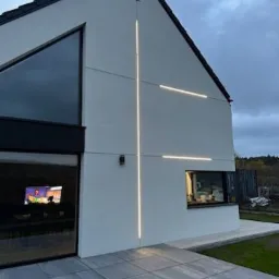 Nowoczesny dom jednorodzinny z białymi ścianami zewnętrznymi i dekoracyjnym oświetleniem elewacyjnym w kształcie krzyża, widok z zewnątrz na szary chodnik i fragment ogrodu.