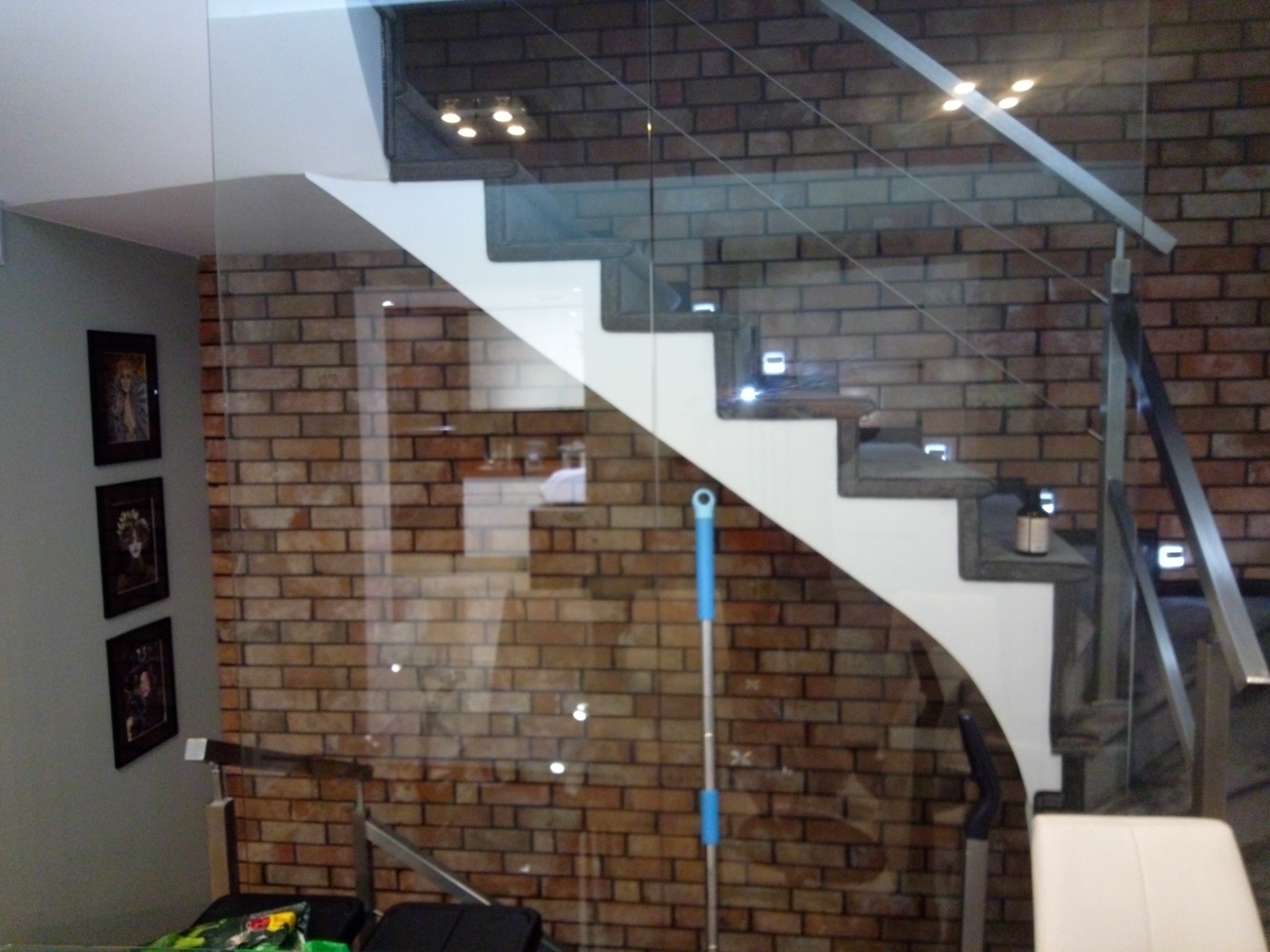 Nowoczesne schody z białymi stopniami i szklanymi balustradami, podświetlone punktowymi lampami, na tle ściany z cegły.