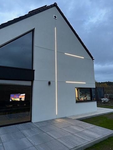 Nowoczesny dom jednorodzinny z białymi ścianami zewnętrznymi i dekoracyjnym oświetleniem elewacyjnym w kształcie krzyża, widok z zewnątrz na szary chodnik i fragment ogrodu.