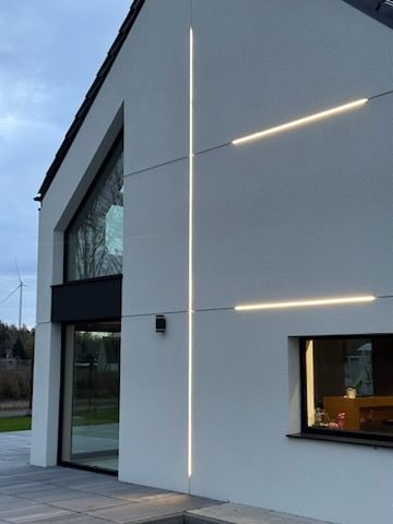 Nowoczesny dom jednorodzinny z białym tynkiem elewacyjnym i dekoracyjnym oświetleniem LED w kształcie linii krzyżujących się na ścianie. Widoczny fragment okna i drzwi balkonowych, a w tle turbina...